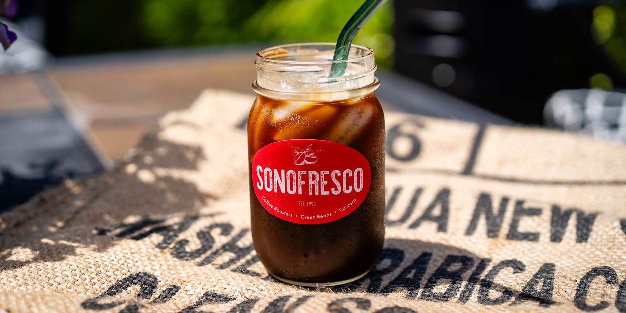 A glass of iced coffee on a green coffee burlap bag under the sunlight, condensation forming on the glass, ice cubes melting, creating a refreshing scene.